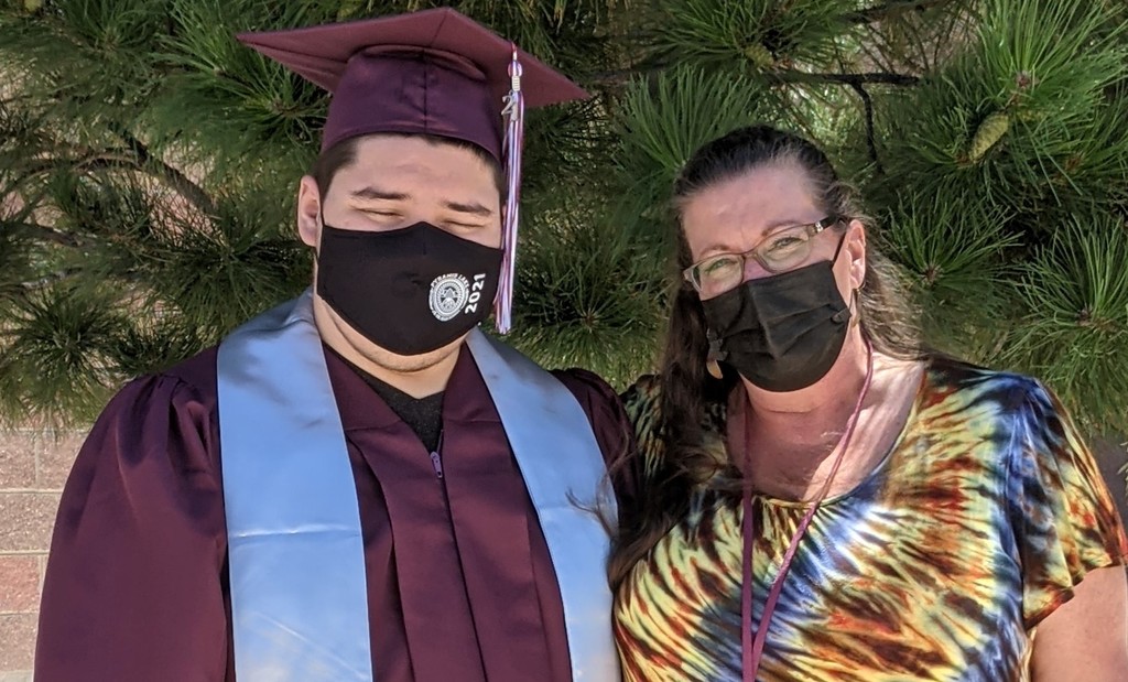 Photo of a graduate in a maroon cap and gown standing with a teacher in a tie dye shirt. They are both wearing masks. 