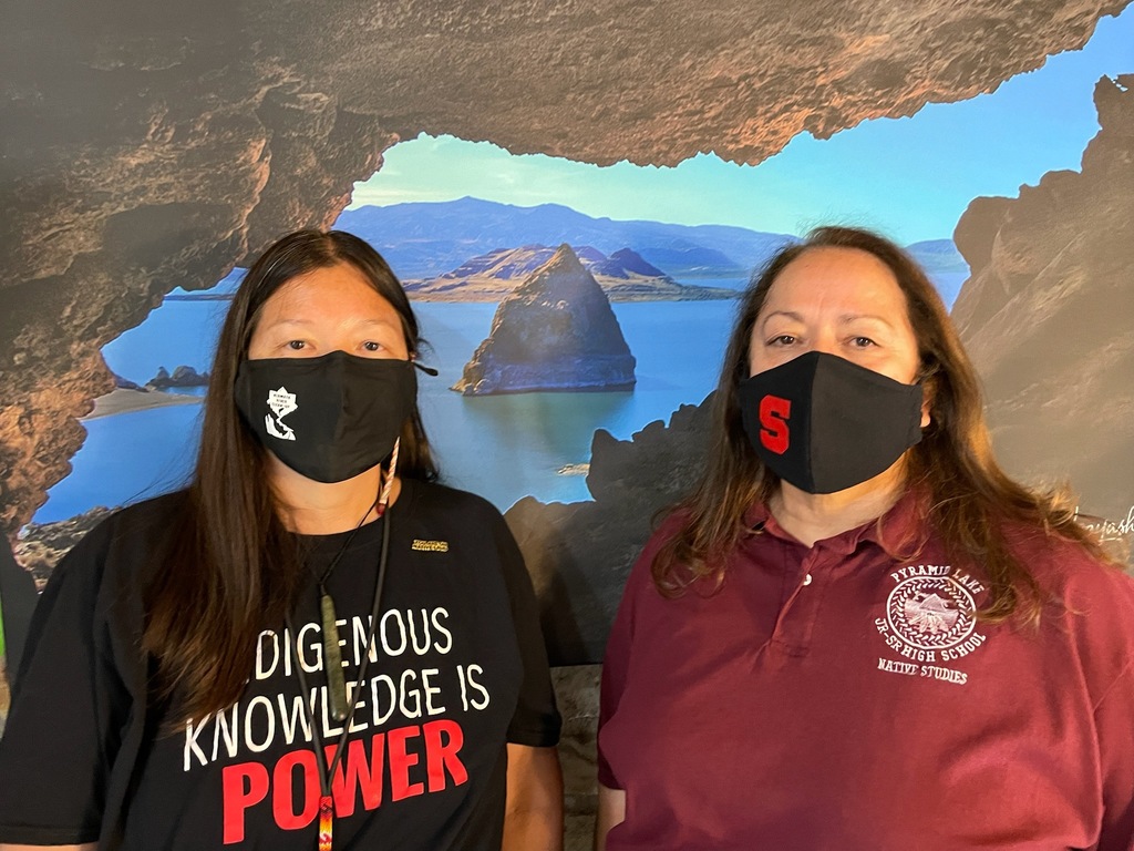 Picture of two women wearing masks, standing in front of a picture on Pyramid Lake and the Pyramid. 